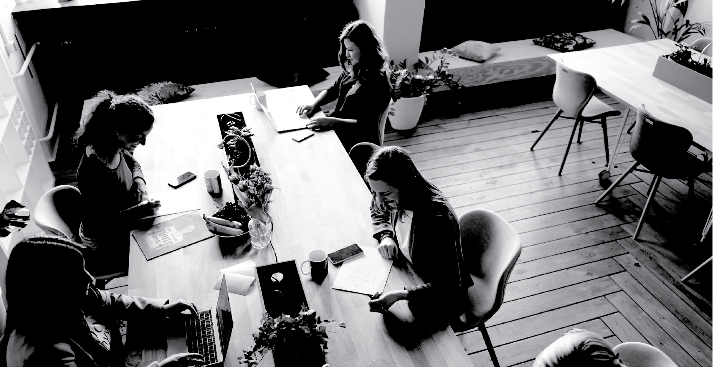 A studio seen from above: people working and laughing around a long wooden table, in black and white
