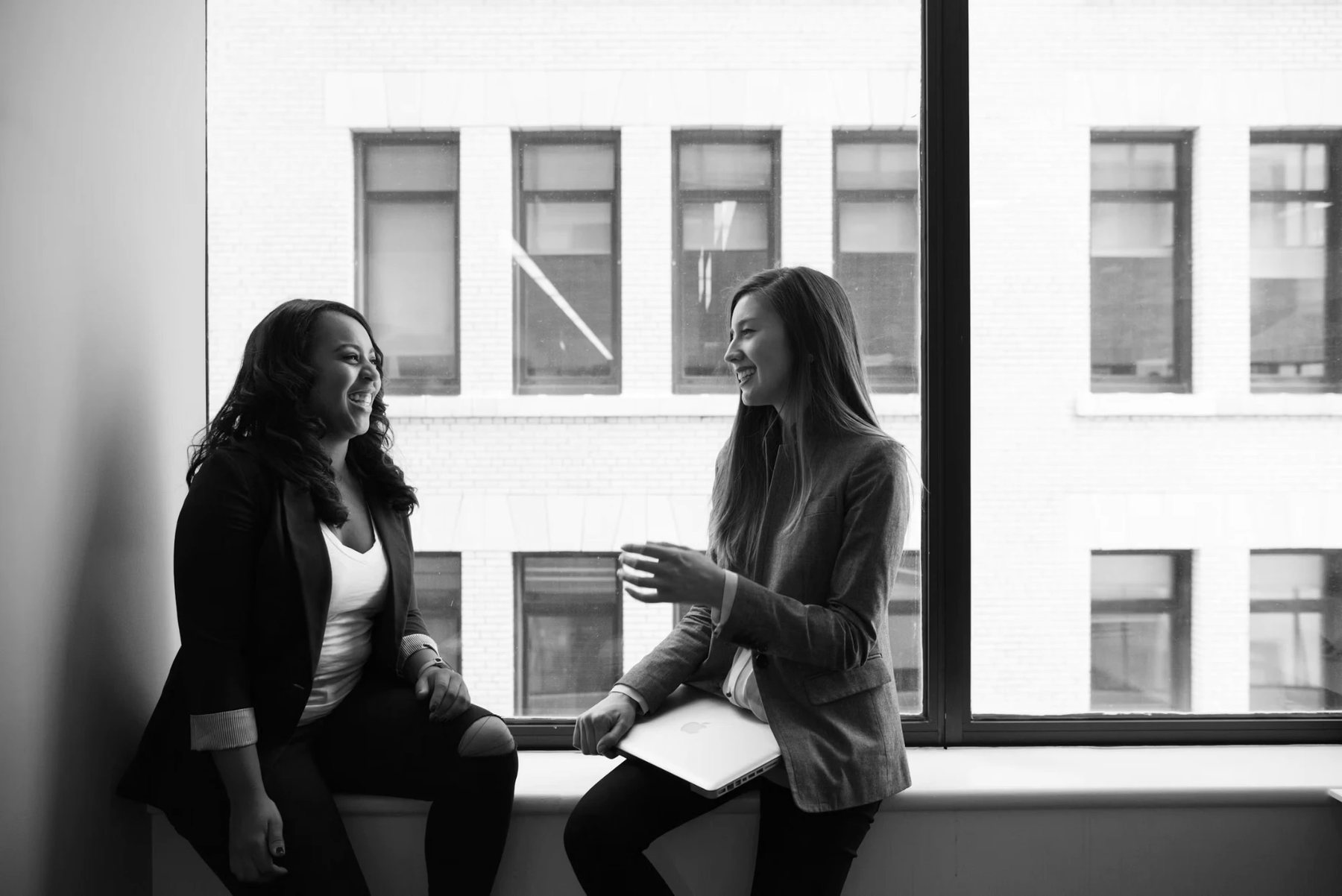 Two women talking and laughing on a sill by a large office window, one holding a laptop